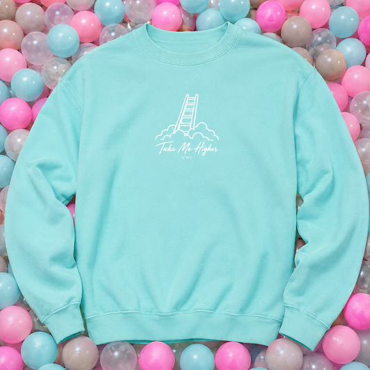 A light aqua crewneck sweatshirt with a white graphic of a ladder climbing out of clouds and the text 'Take Me Higher' on the chest, laid flat on a bed of pink, blue, and purple plastic balls.