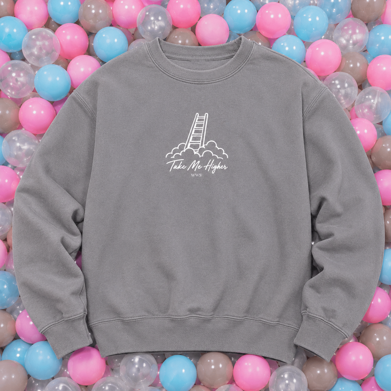 A gray crewneck sweatshirt with a white graphic of a ladder climbing clouds and the text 'Take Me Higher' and '2025' on the chest, displayed on a bed of pink, blue, and clear plastic balls.