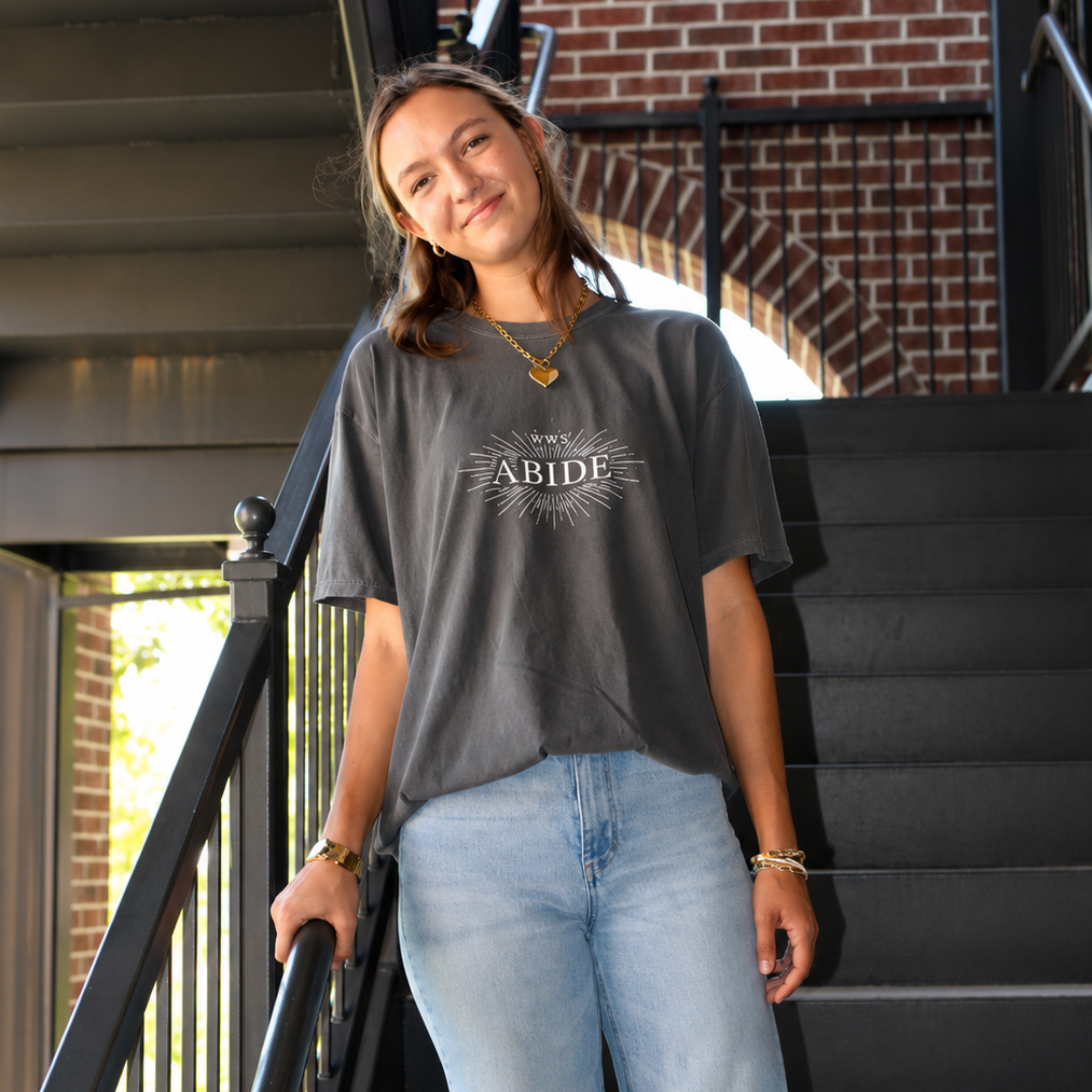 A woman stands on an outdoor staircase wearing a dark charcoal gray graphic t-shirt with 'ABIDE' printed on the chest, paired with light wash blue jeans.