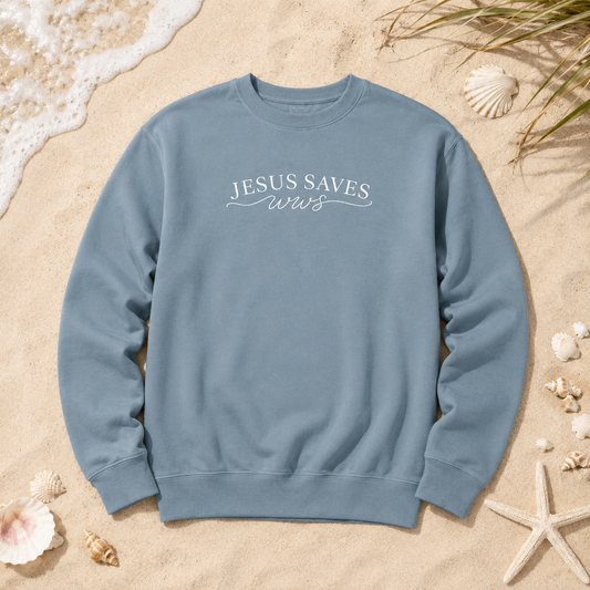 A dusty blue crewneck sweatshirt with 'JESUS SAVES us' printed in white across the chest, laid flat on a sandy beach styled with seashells and starfish.