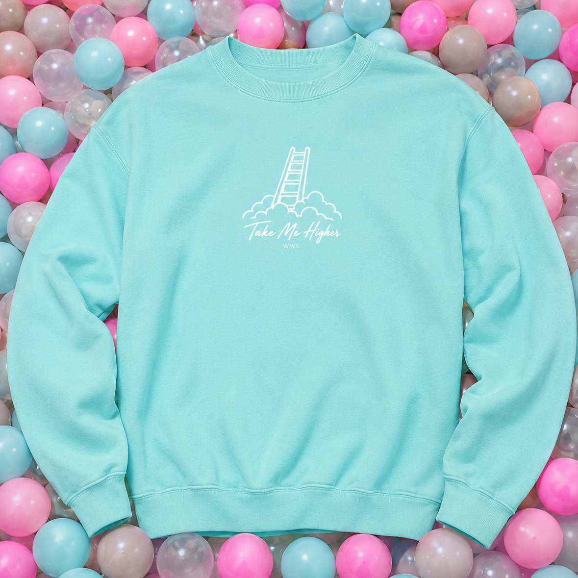 A light aqua crewneck sweatshirt with a white graphic of a ladder climbing out of clouds and the text 'Take Me Higher' on the chest, laid flat on a bed of pink, blue, and purple plastic balls.
