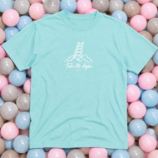A mint green short-sleeve t-shirt with a white graphic of a ladder rising through clouds and the text 'Take Me Higher' printed on the chest. The shirt is laid flat on a bed of pastel pink, blue, and gray balls.