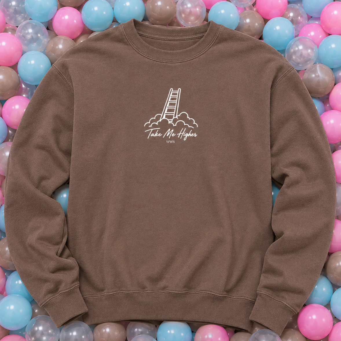 A brown crewneck sweatshirt with a white graphic of a ladder climbing mountains and the text 'Take Mr. Hedges' on the chest, laid flat on a bed of pink, blue, and clear plastic balls.