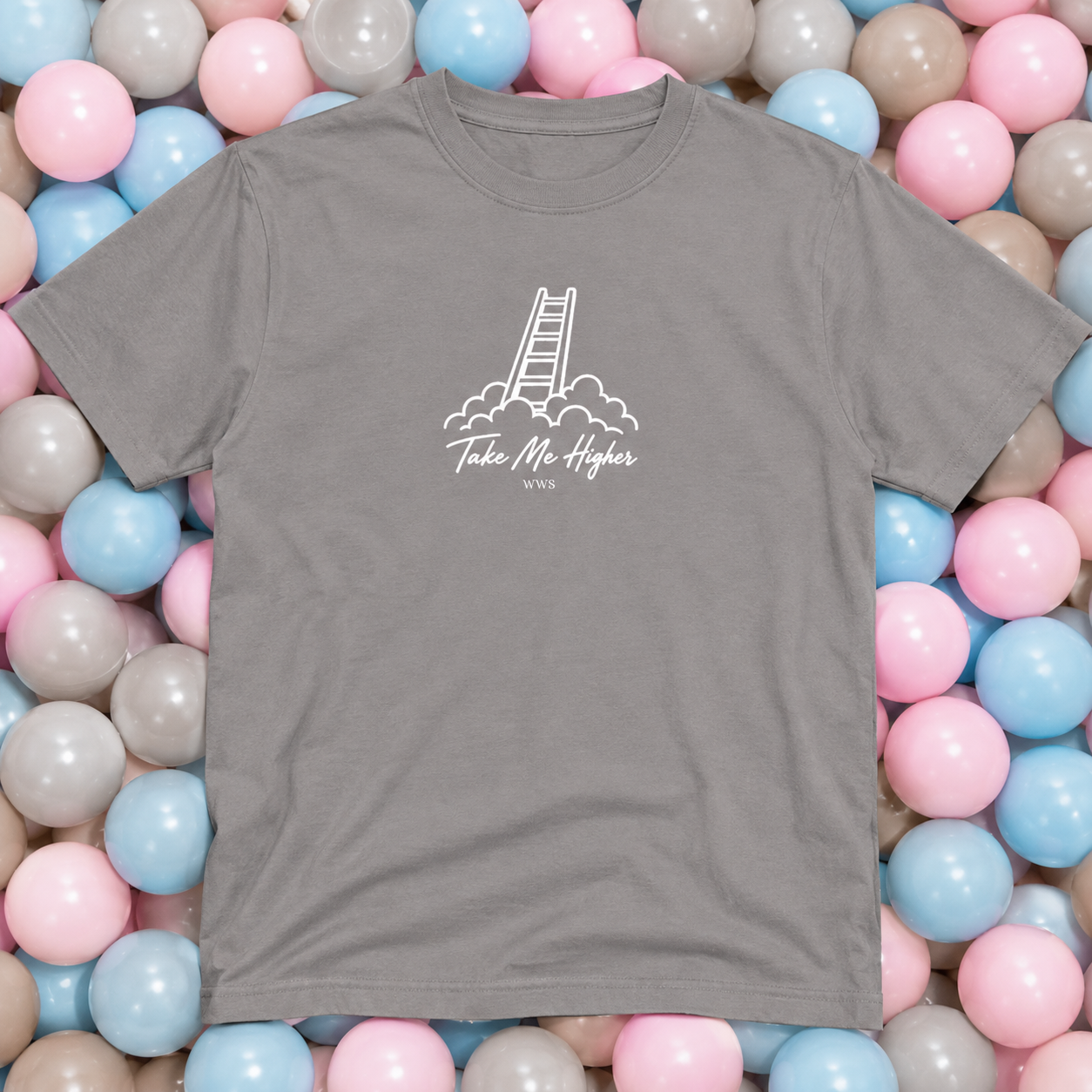 A gray short-sleeve t-shirt with a white graphic of a ladder and the text 'Take Me Higher' laid flat on a bed of pastel pink, blue, and gray plastic balls.