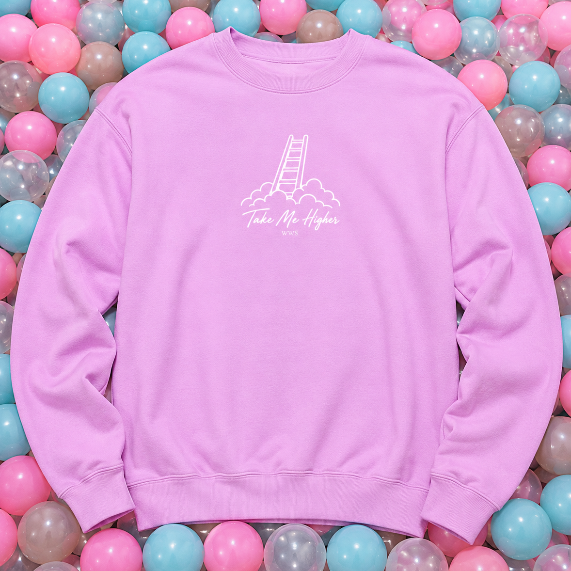 A light purple crewneck sweatshirt with a white graphic of a ladder in the clouds and the text 'Take Me Higher' on the chest, laid flat on a bed of pink, blue, and clear plastic balls.