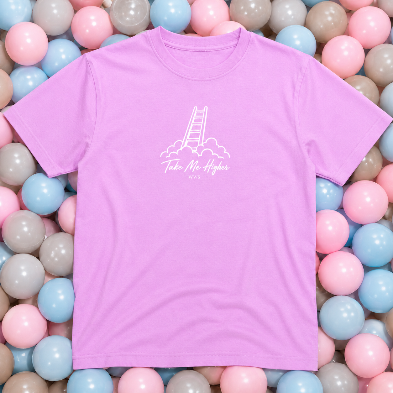A light purple short-sleeve t-shirt with a white graphic of a ladder climbing out of clouds and the text 'Take Me Higher' printed on the chest. The shirt is laid flat on a bed of pastel pink, blue, and gray plastic balls.