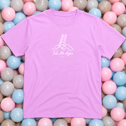 A light purple short-sleeve t-shirt with a white graphic of a ladder climbing out of clouds and the text 'Take Me Higher' printed on the chest. The shirt is laid flat on a bed of pastel pink, blue, and gray plastic balls.